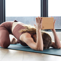 Person on mat in Childs Pose with the Performance Cork Block in hands with arms bent at the elbow - block above back of head. 